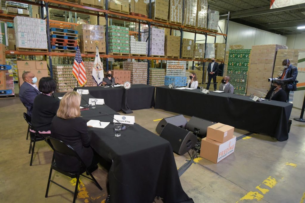 A photo of the interior of the Feeding Northeast Florida warehouse. There are 5 tables arranged in an upside-down U formation, with Vice President Harris at the head of the table. From bottom left to right, Susan King, Audrey Gibson, Al Lawson, Vice President Harris, Nikki Fried, Garrett Dennis, and Dr. Diana Greene are pictured.